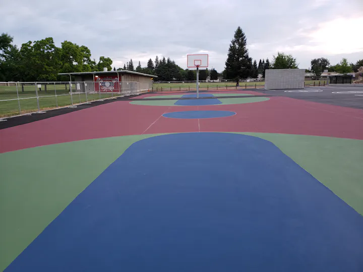 A colorful outdoor basketball court with various painted sections, a hoop, and a nearby building in a park setting.