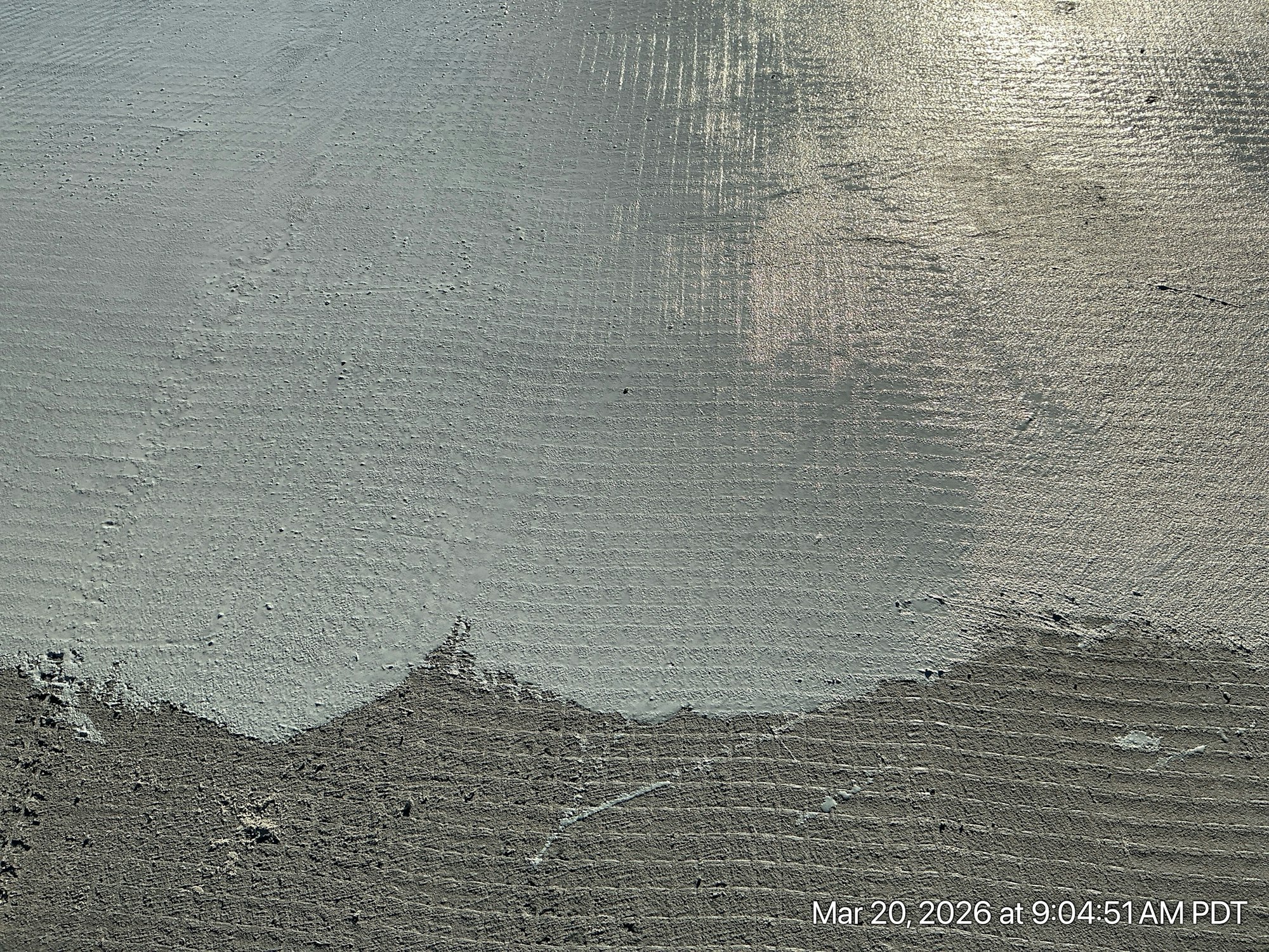 The image shows a textured surface, possibly wet concrete or sand, with variations in color and light reflections.