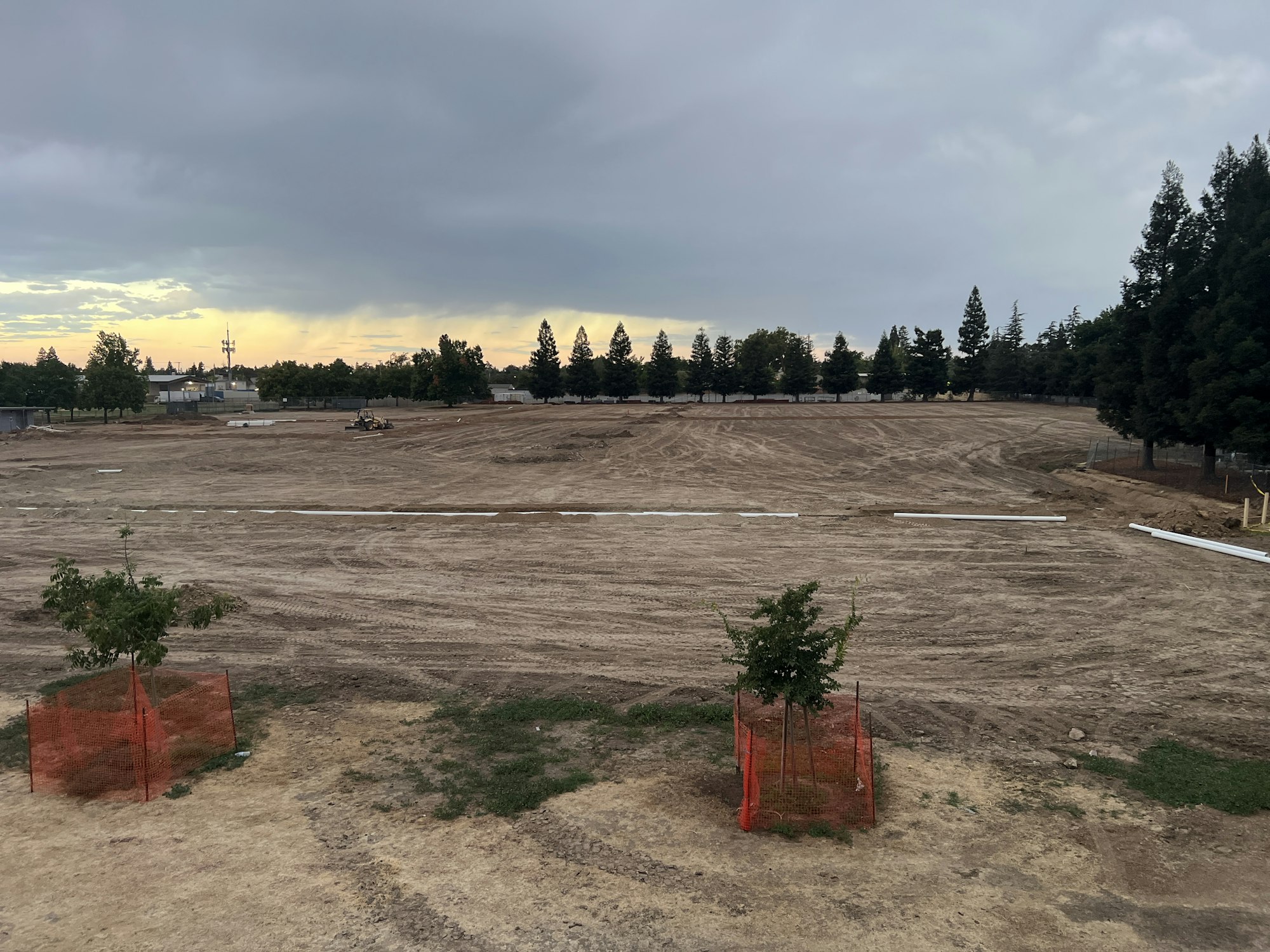 A large dirt field with some scattered trees and orange fencing under a cloudy sky.