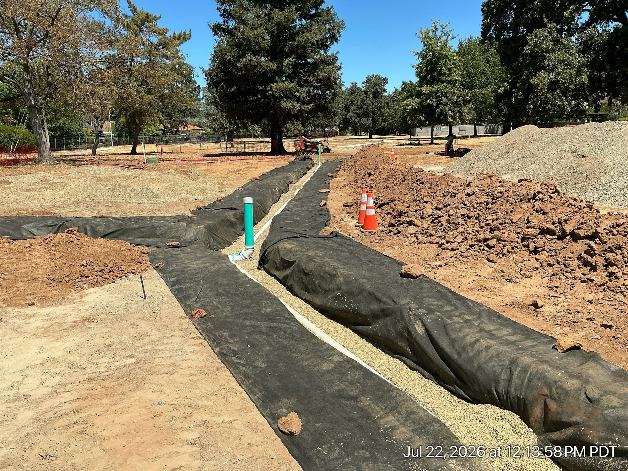 The image shows a construction site with excavated soil, lined areas, drainage pipes, and gravel preparation under a clear blue sky.