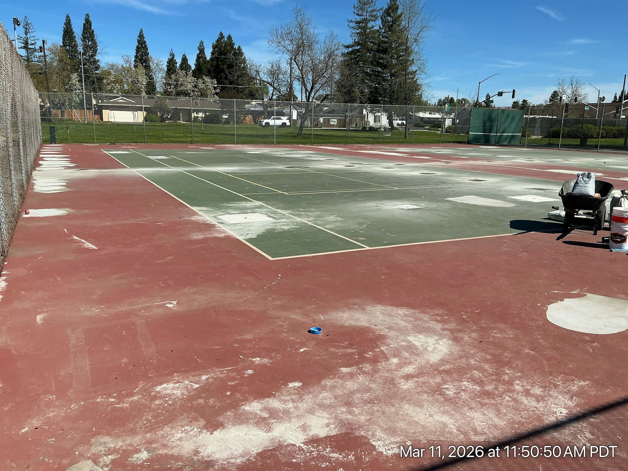 The image shows a partially maintained tennis court with red and green surfaces, surrounded by a chain-link fence and trees.