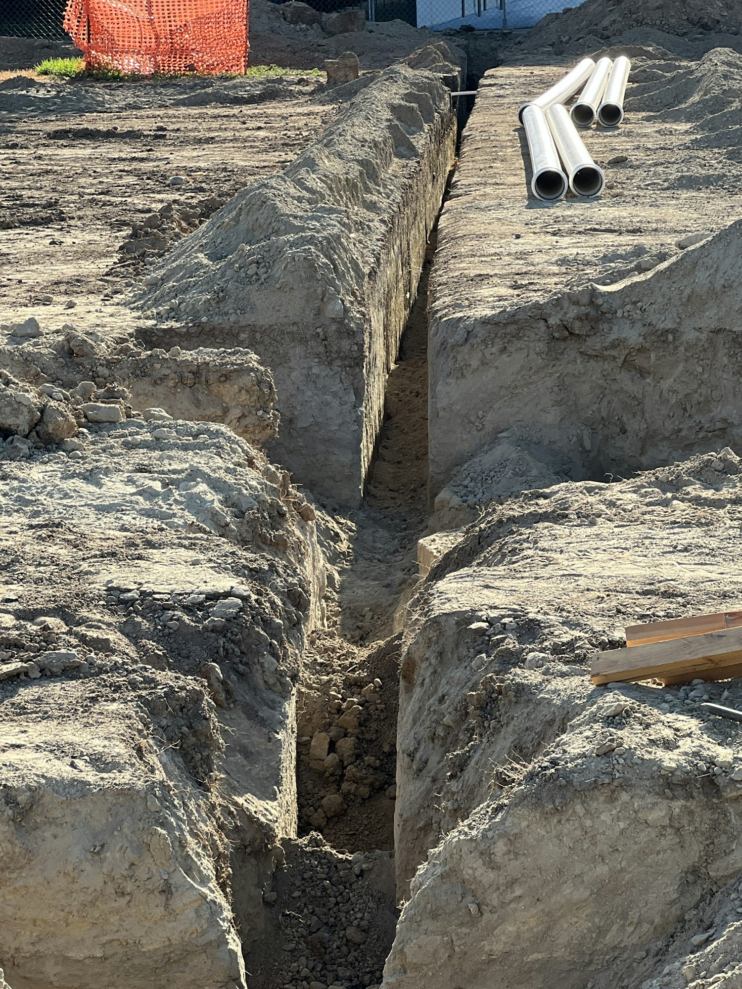A trench dug in the ground with several metal pipes lying on the surface nearby.