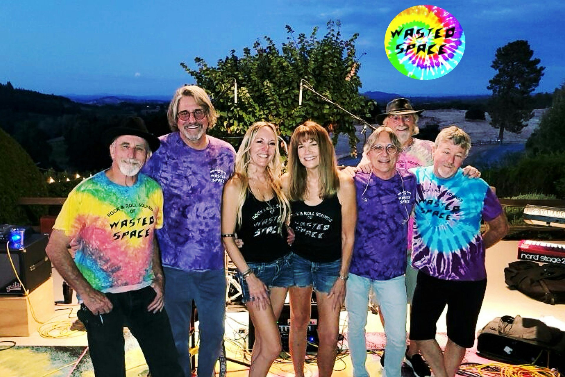 A group of people in colorful tie-dye shirts labeled "Wasted Space," posing outdoors with musical equipment.