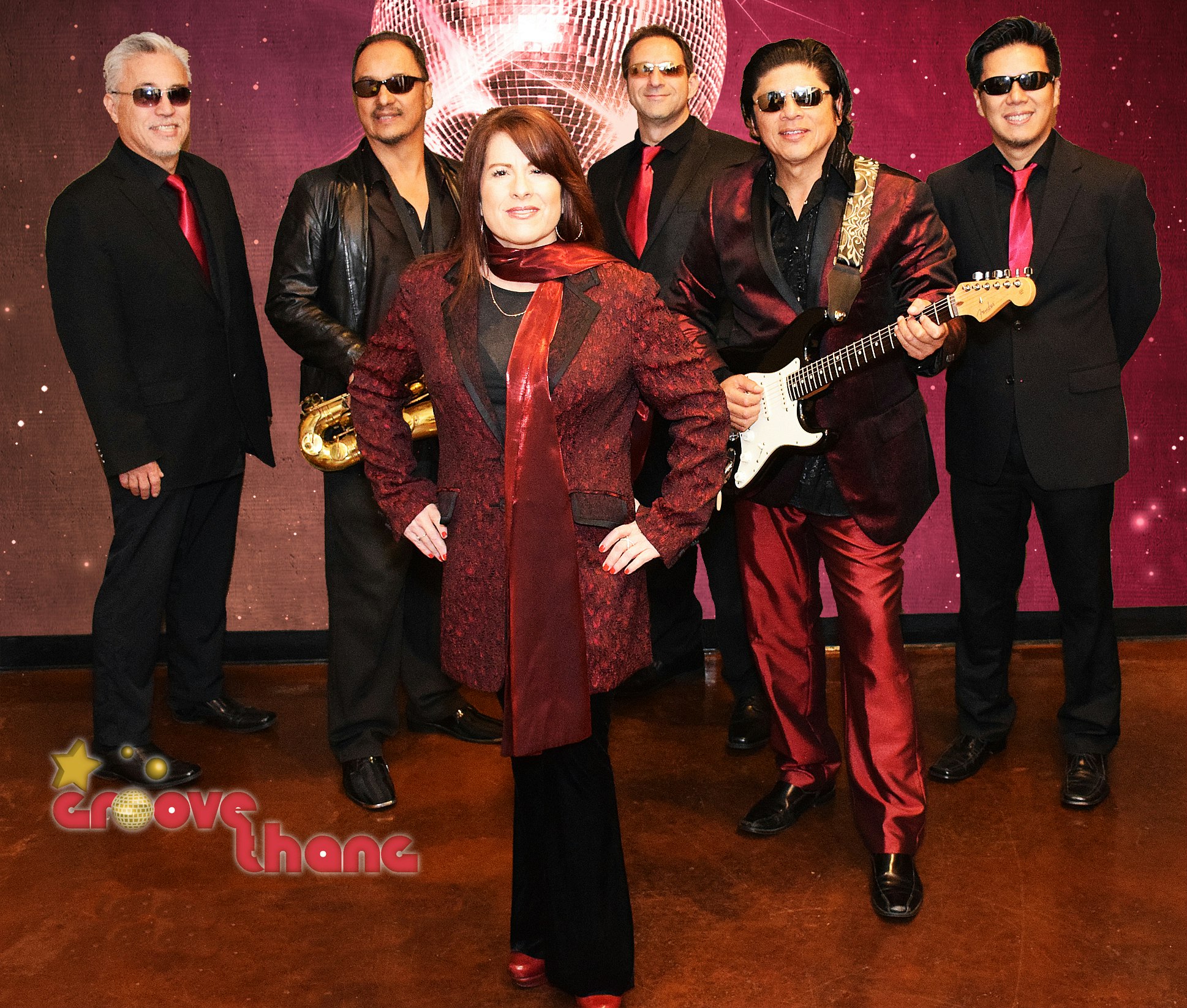 A band of six people posing together; one holds a guitar, another a saxophone. They're dressed in black and red attire with sunglasses.