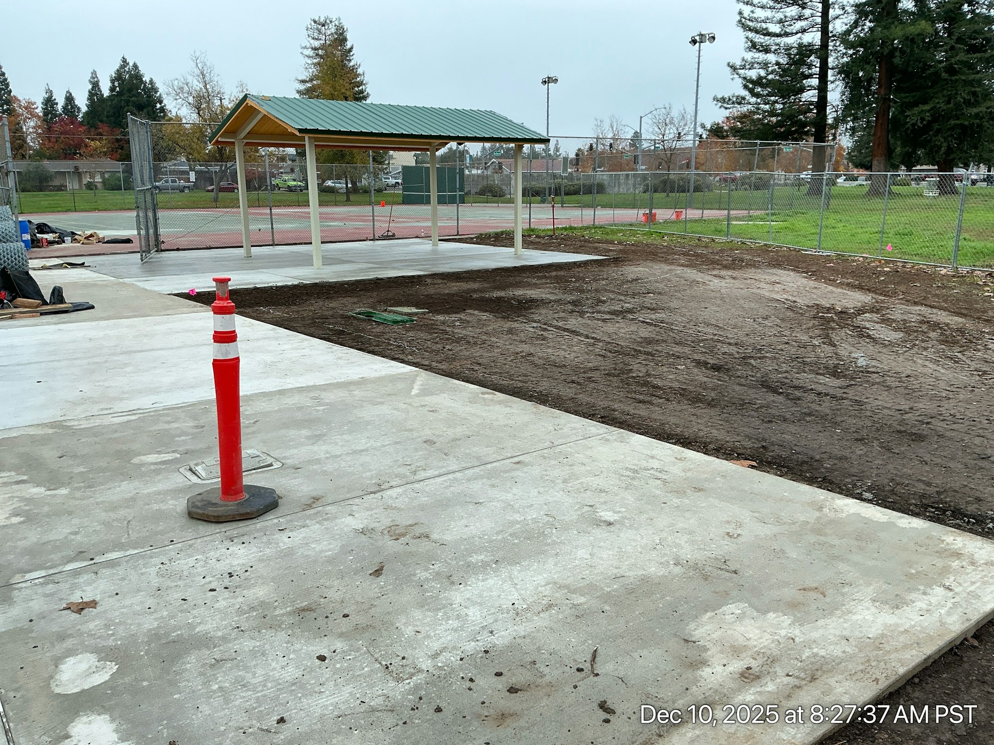 The image shows a partially constructed area in a park, featuring a shelter, concrete pathways, and a dirt patch nearby.