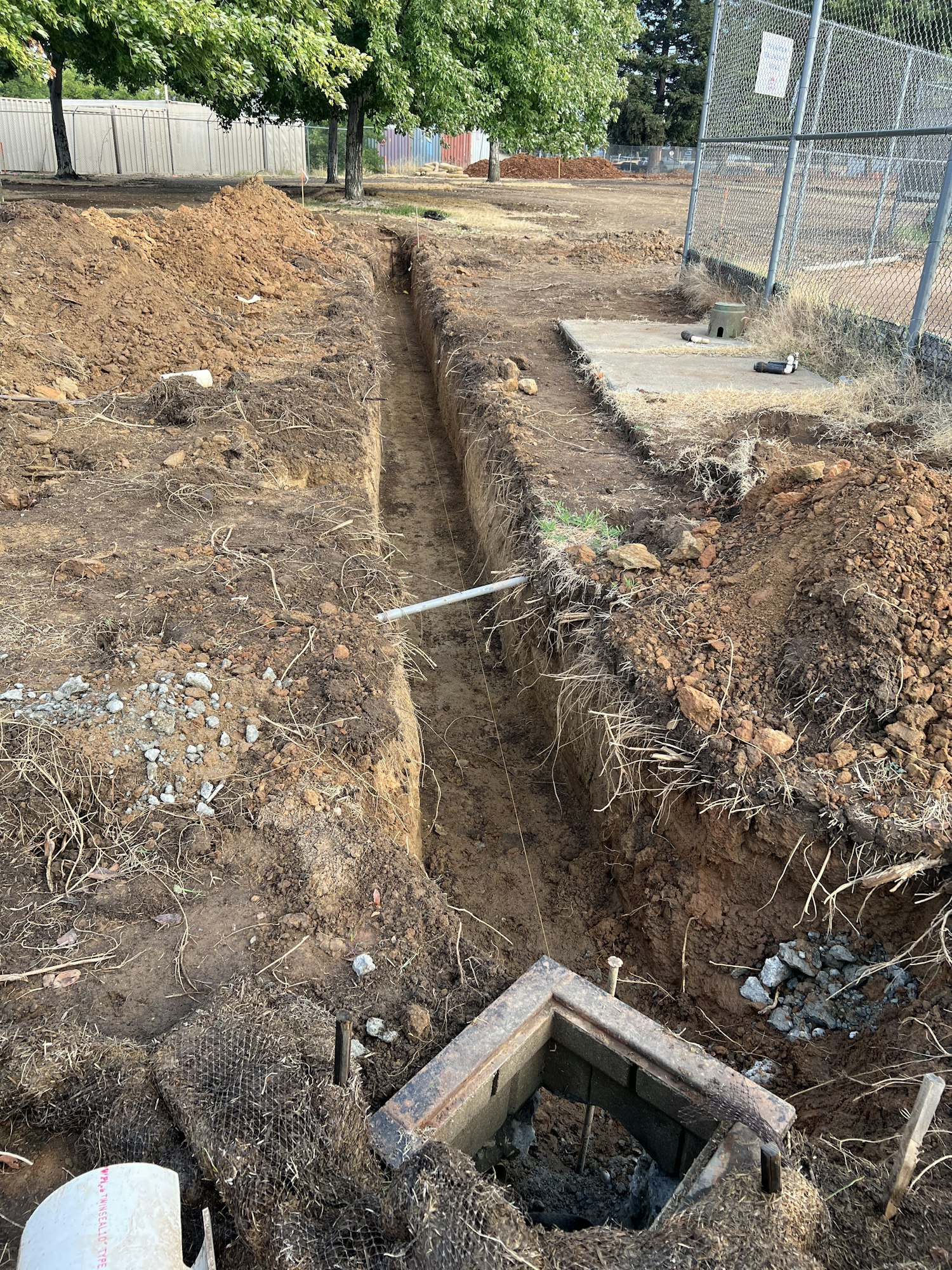 A narrow trench dug into the ground, possibly for construction or piping, next to a fence and trees.