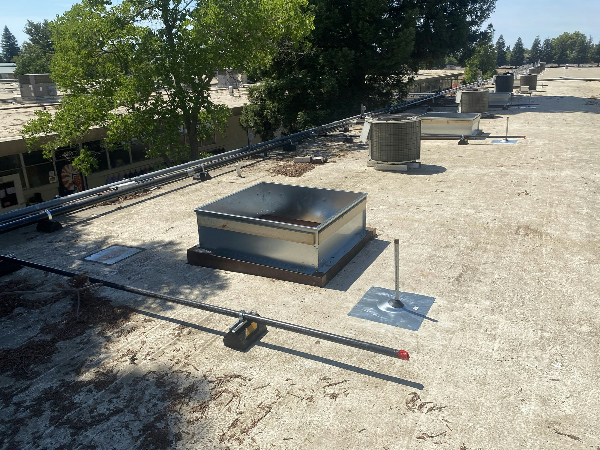 A flat rooftop with HVAC units, pipes, and trees in the background.
