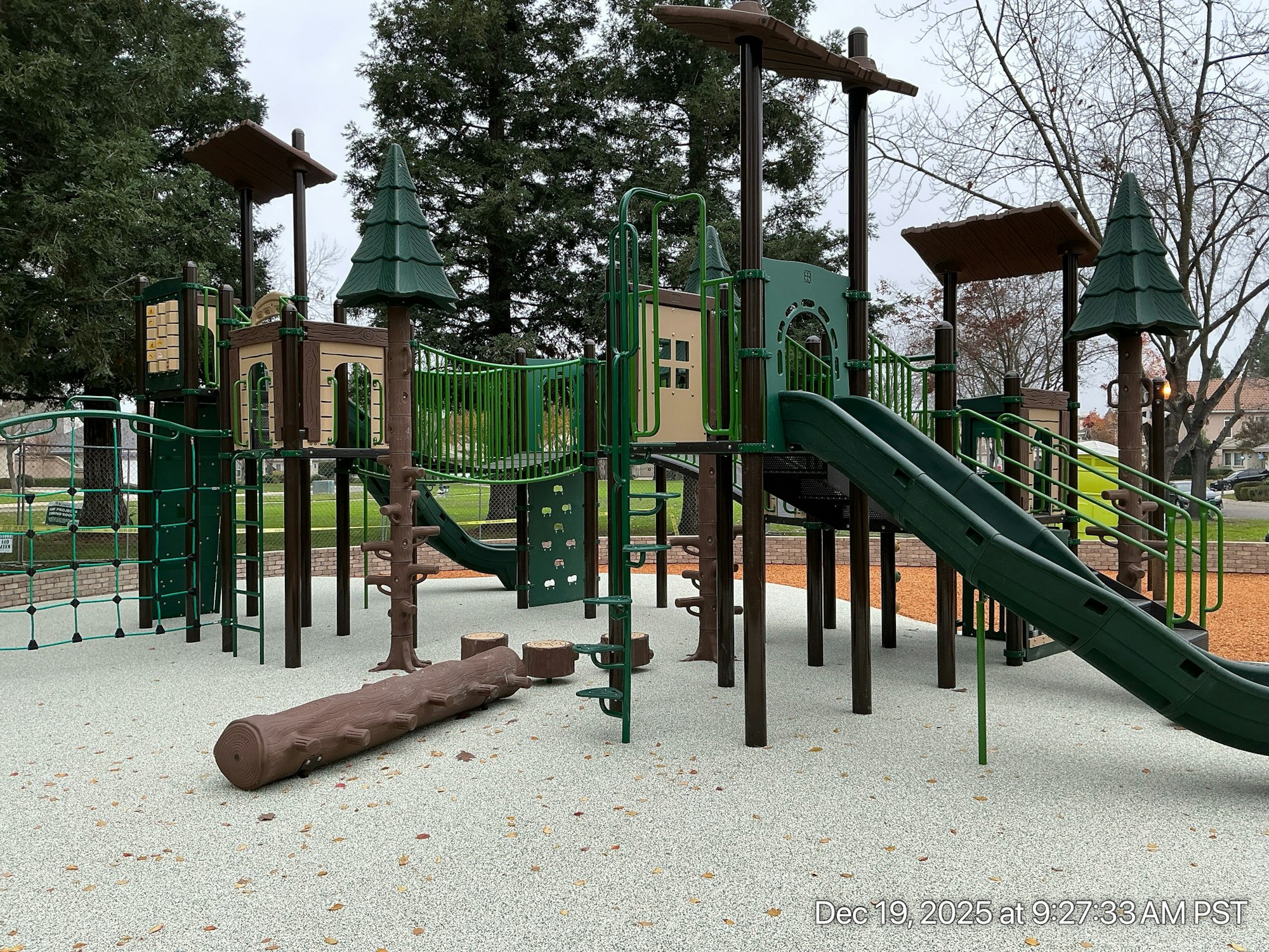A playground with green slides, climbing structures, and a log feature on a soft surface, surrounded by trees.