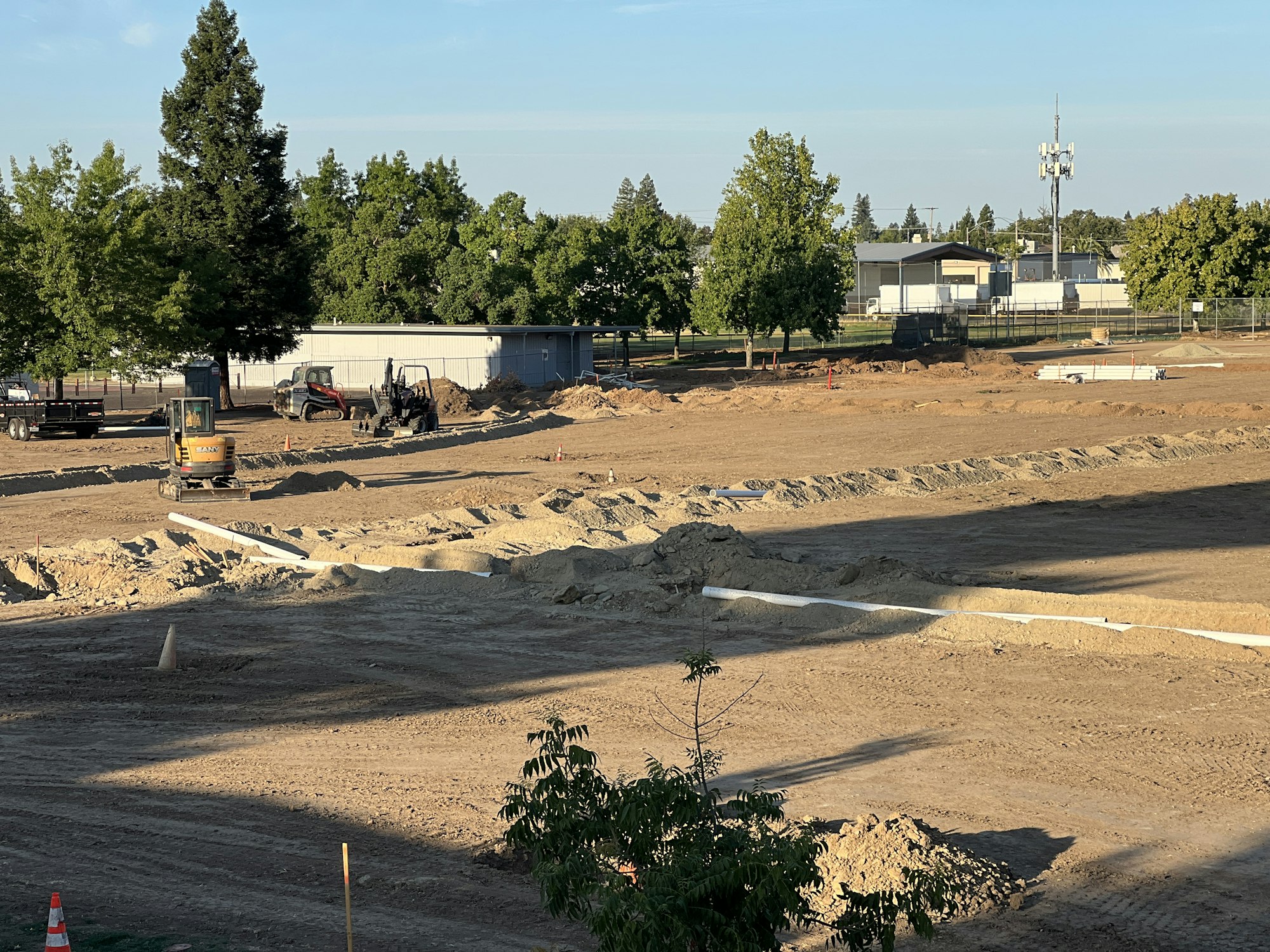 A construction site with earth movers, pipes, and piles of dirt under a clear sky, surrounded by trees.