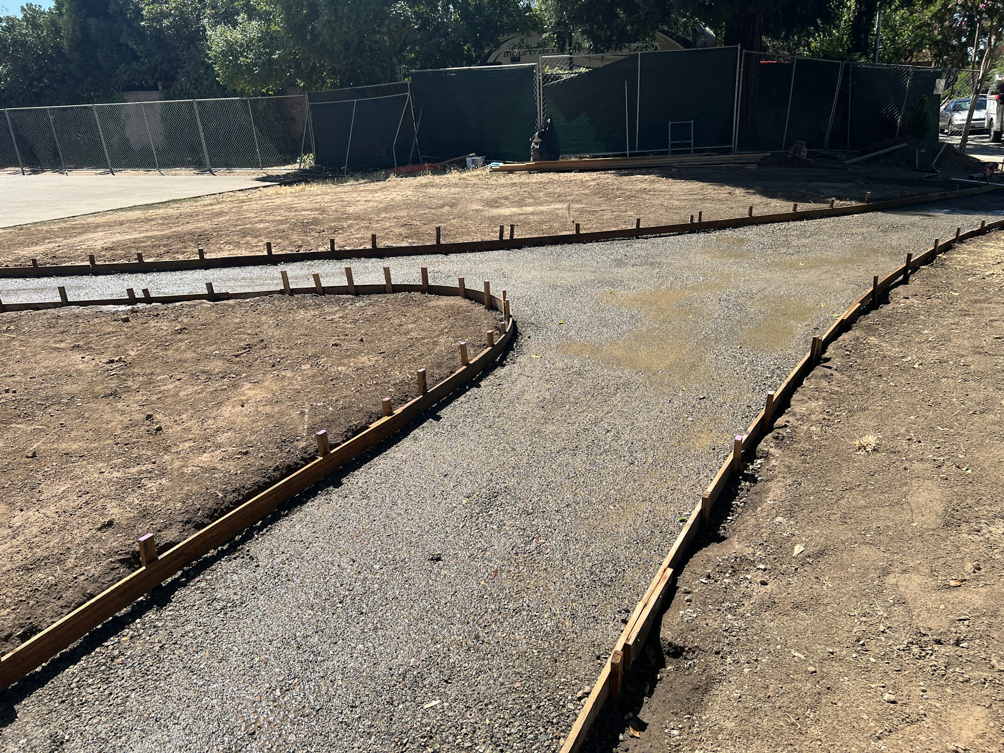 Curved path under construction with wooden forms and gravel, surrounded by dry ground and fence.