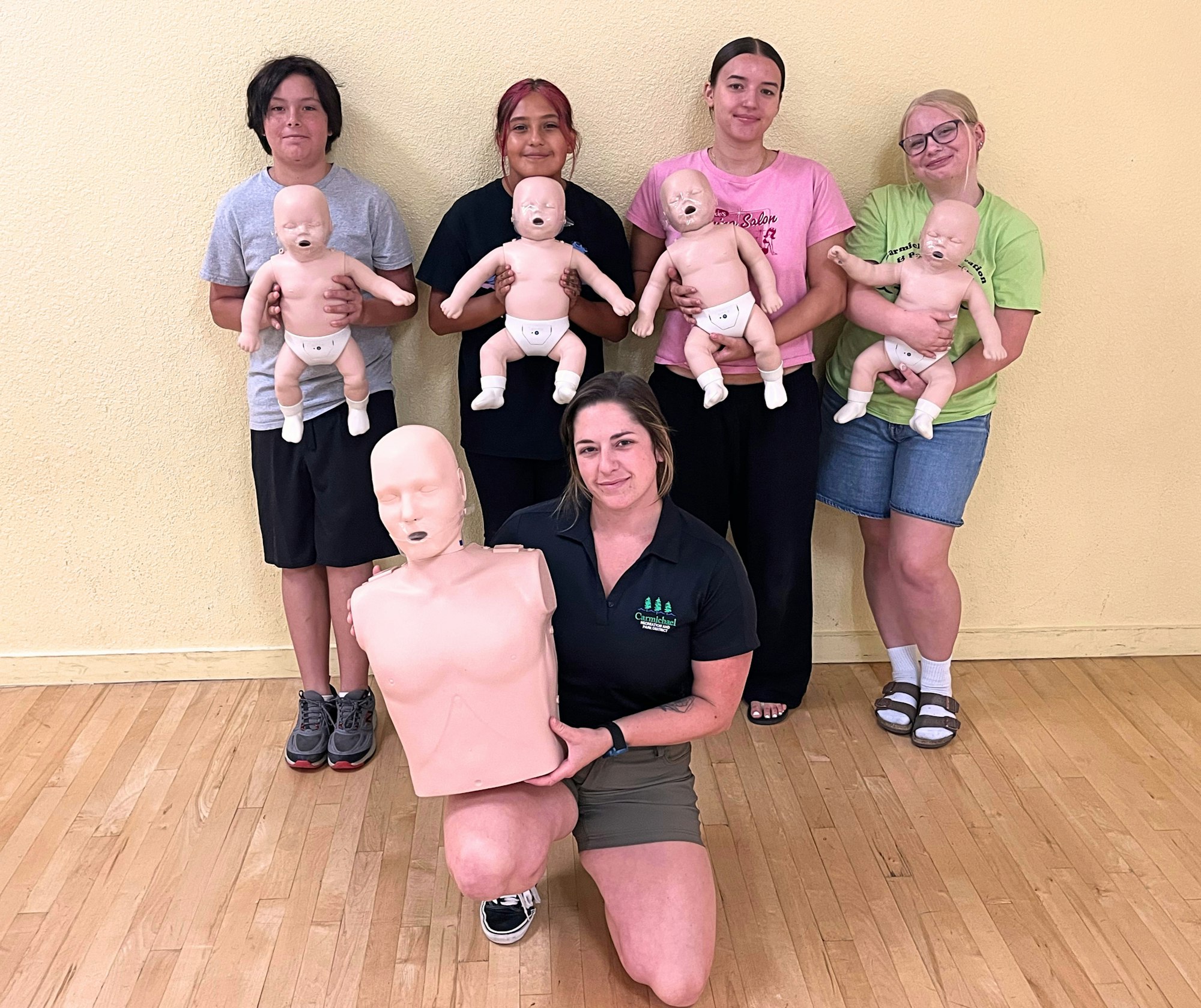 A group of five people poses with training mannequins (infant and adult) in a classroom setting.