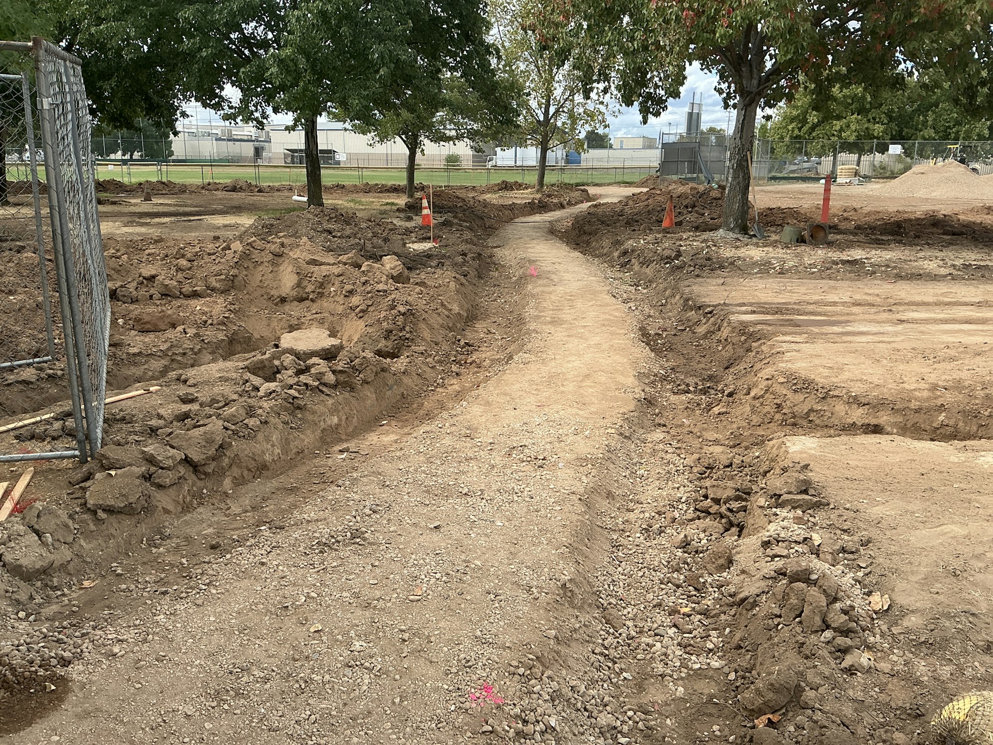 A dirt path in a construction site surrounded by trees, with some trenches and orange cones.