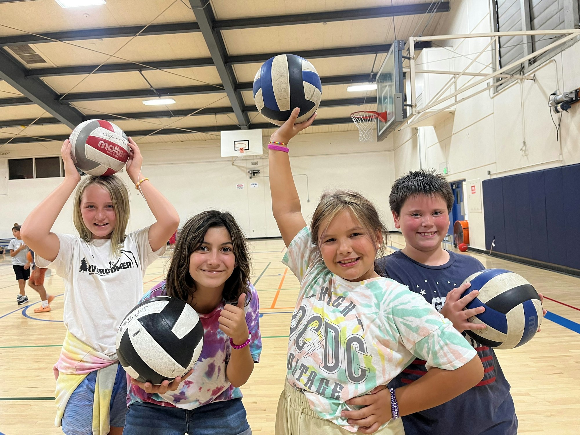 Four children are posing with volleyballs in a gym, smiling and having fun during an activity or game.