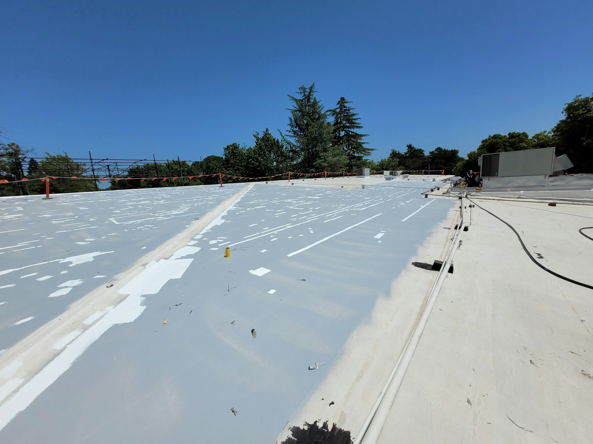 A large, flat roof with patchy white coating, various vents, pipes, and a safety barrier at the edge.
