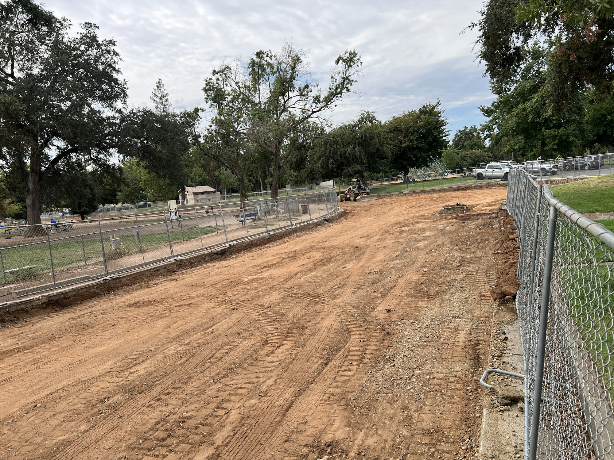 A construction or excavation site with dirt, fencing, and machinery, surrounded by trees and parked vehicles.