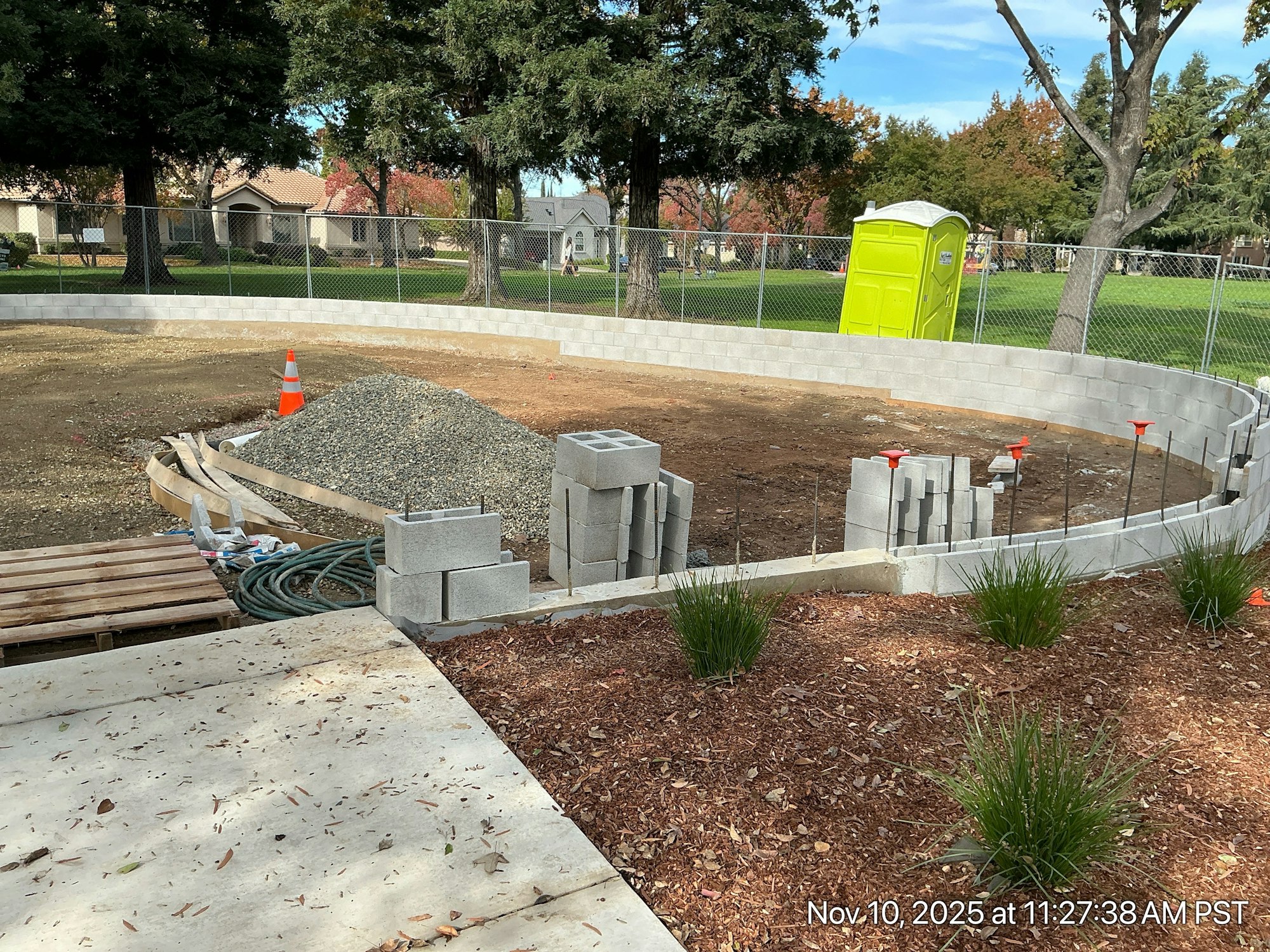 A construction site with a circular wall made of blocks, gravel pile, tools, and a portable restroom in the background.