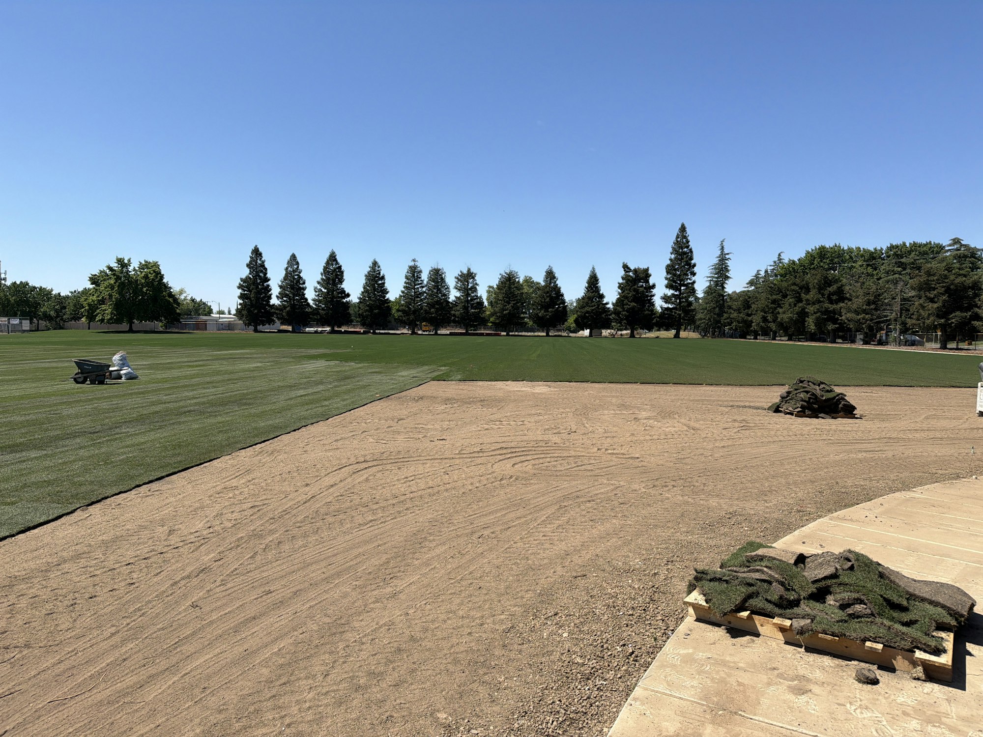 A landscaped area with freshly laid grass, piles of turf, and a wheelbarrow against a clear blue sky and trees in the background.