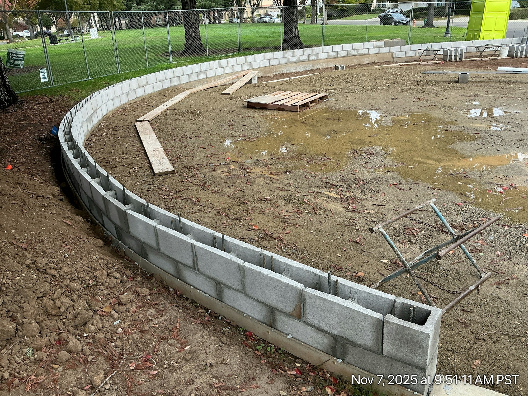 A curved concrete block wall is partially built on a construction site with wooden planks and some water pooling on the ground.