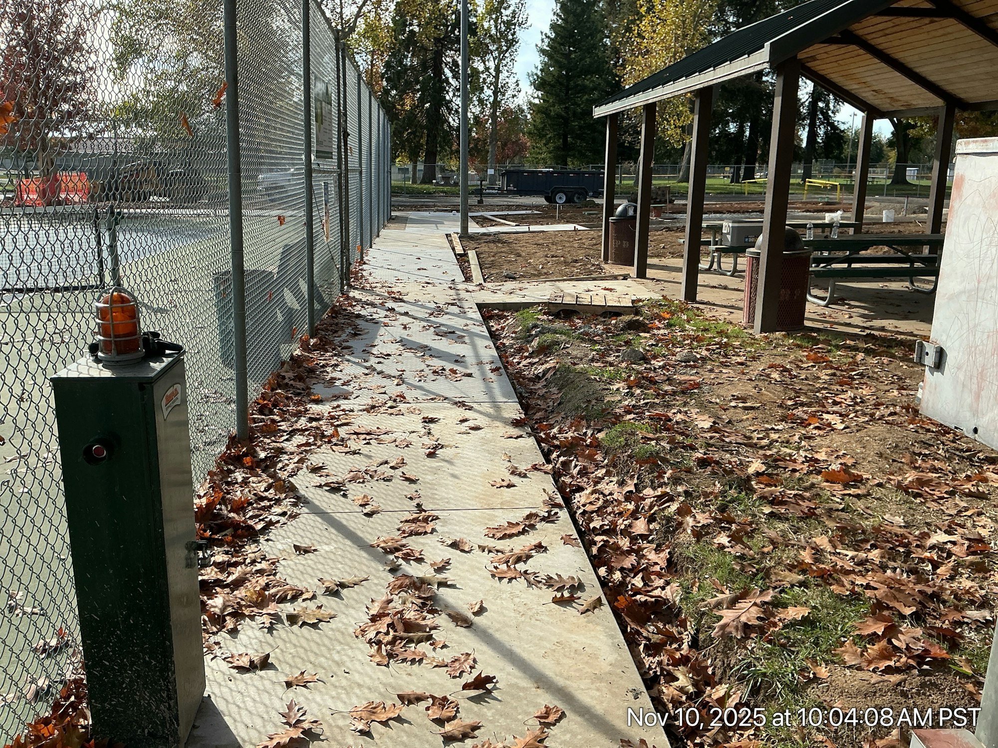 A paved path lined with fallen leaves, adjacent to a fence, leads to a covered picnic area in a park setting.
