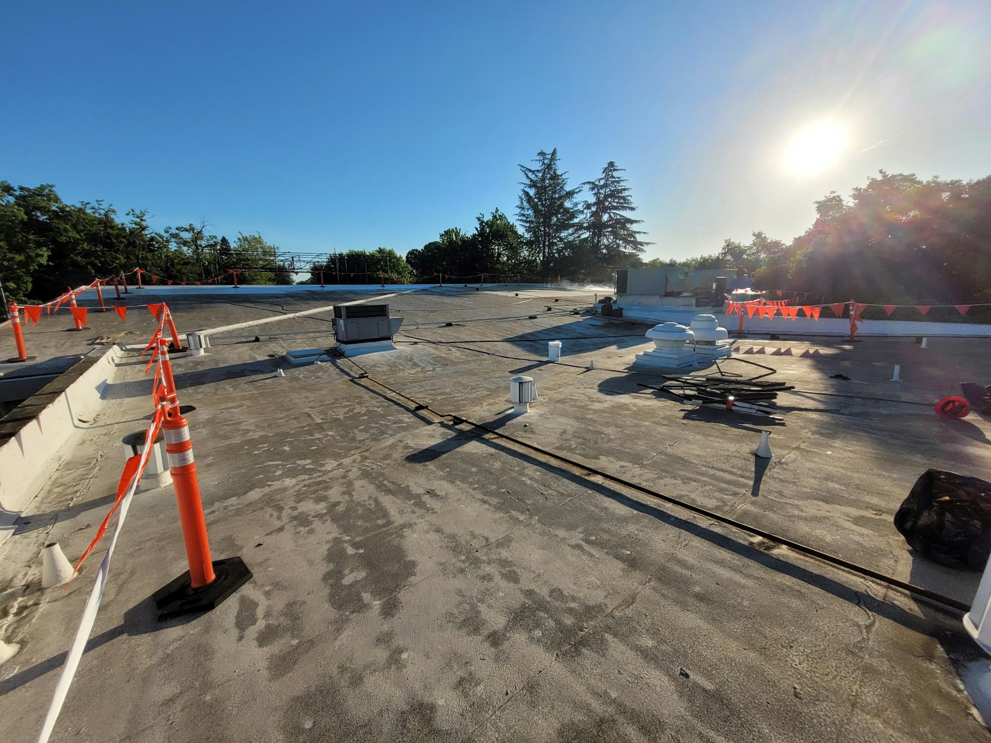 A flat building rooftop with safety cones, vent pipes, and ductwork, bathed in sunlight with trees in the background.