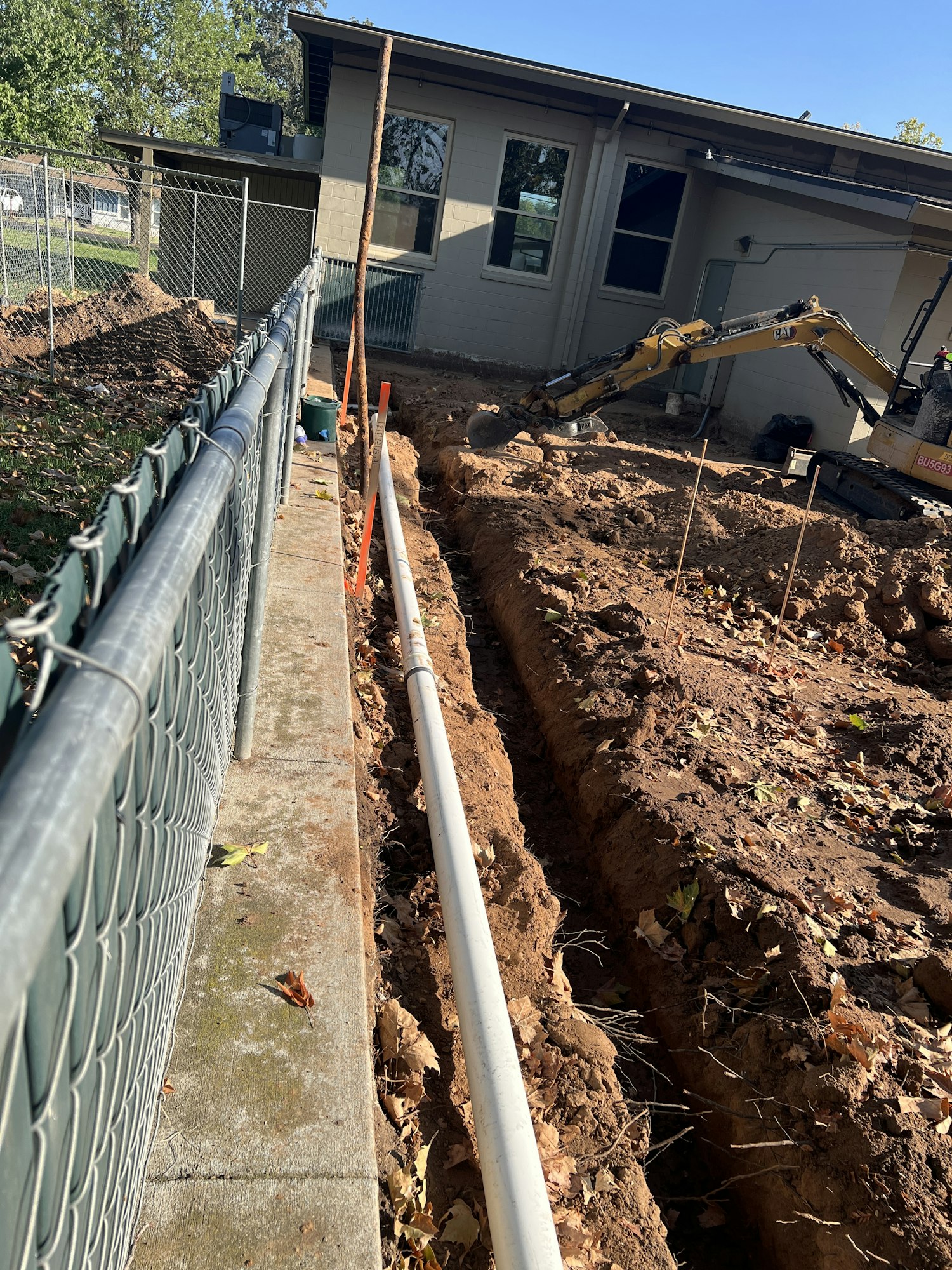A trench with a pipe installation next to a building, a fence, and construction equipment.