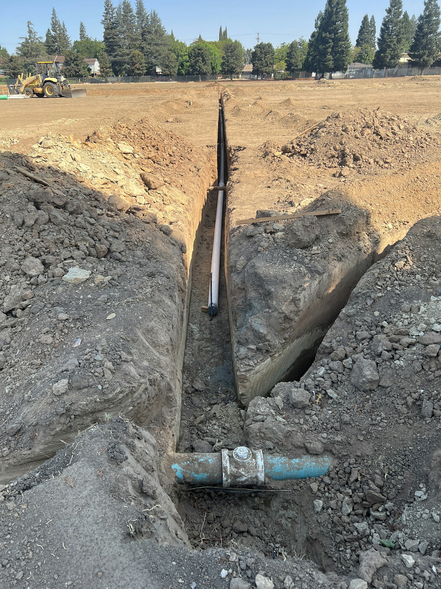 A construction site with a trench containing a pipe, surrounded by dug-up earth and a distant bulldozer.