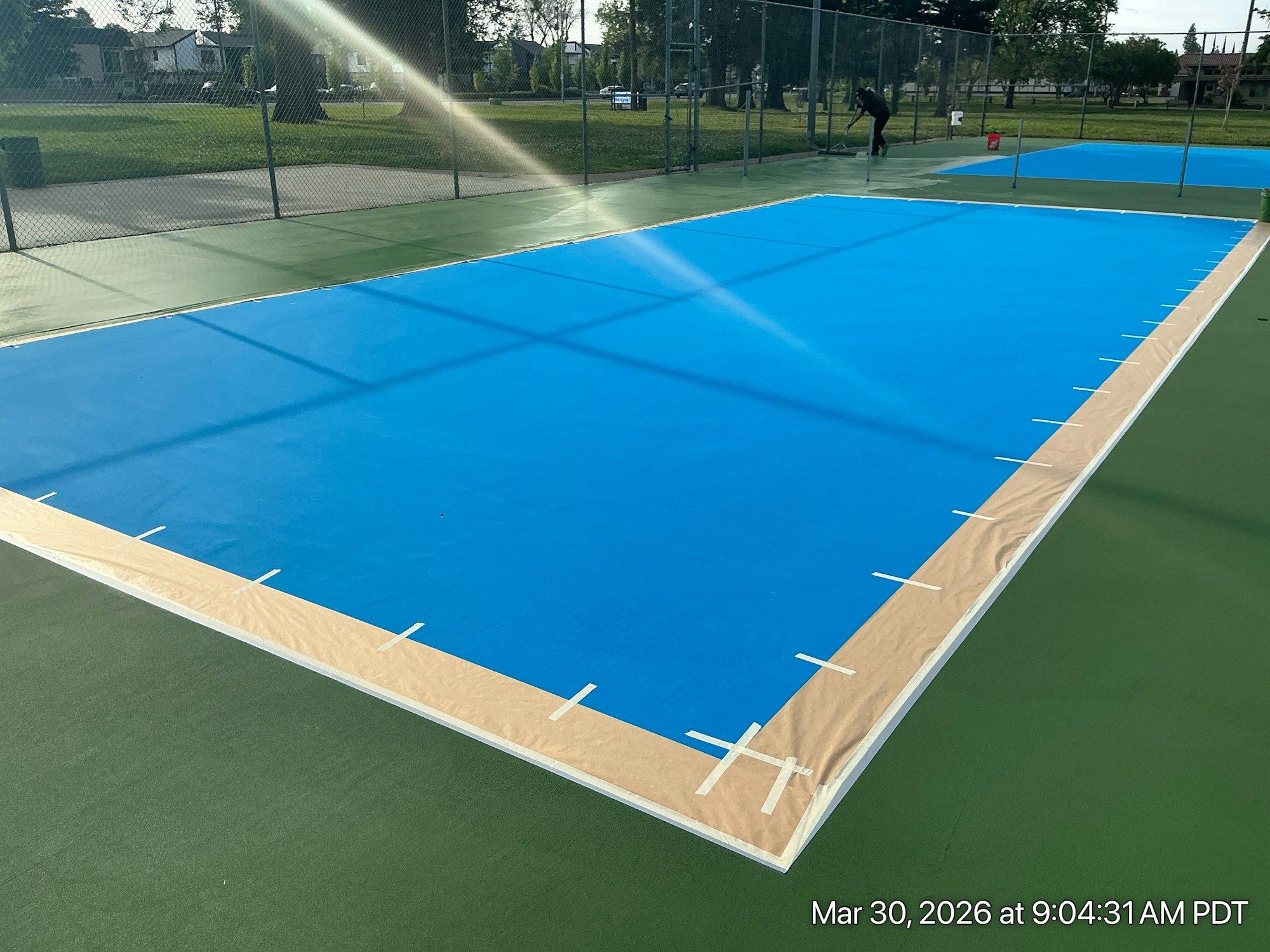 The image shows a blue surface being prepared on a tennis court, with a fence and green surrounding areas visible.