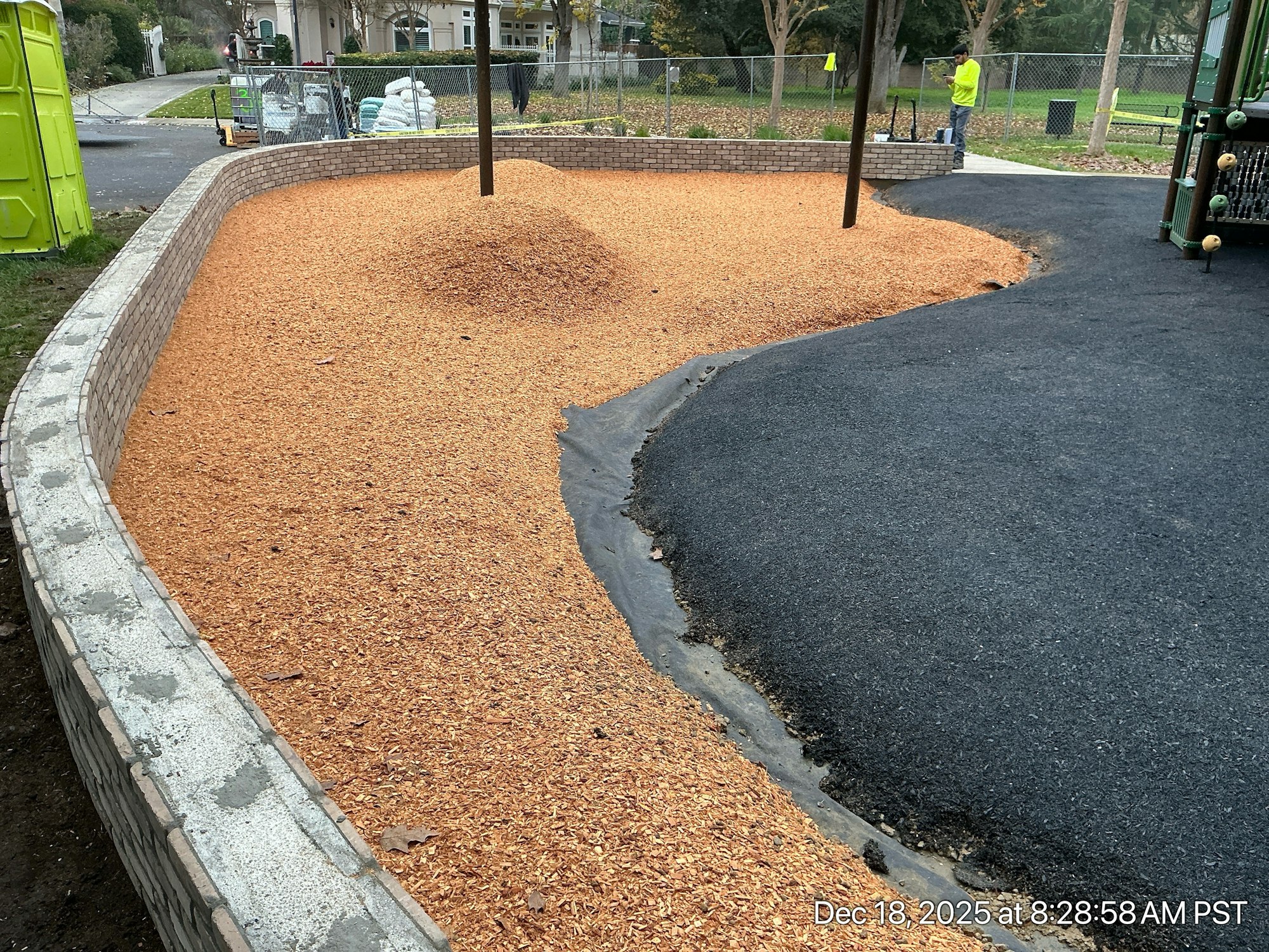 The image shows a playground area with wood chips, a circular wall, and a paved path, with construction workers in the background.