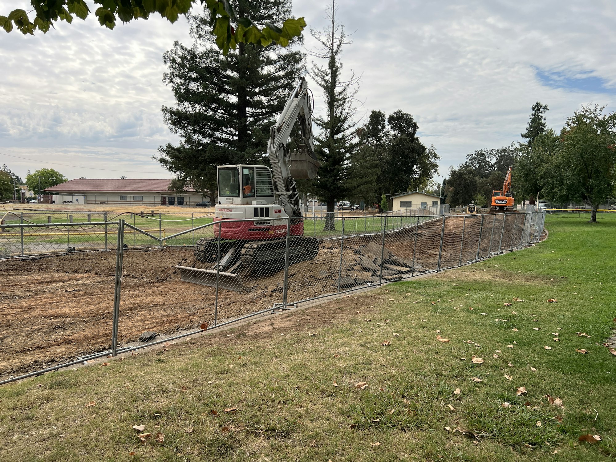 A construction site with excavators, fenced off, surrounded by grass and trees.