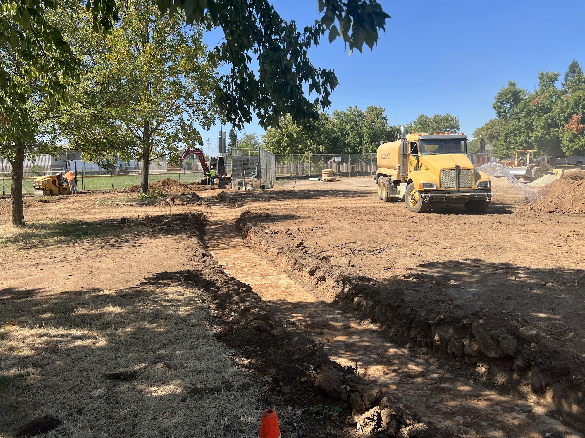 Construction site with a yellow truck, an excavator, and workers. Trees border the area under a clear blue sky.