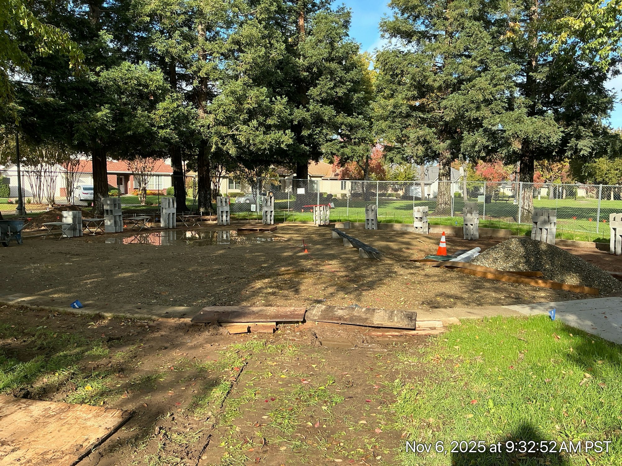 A construction site in a park with scattered materials, concrete blocks, and a gravel pile amid trees.