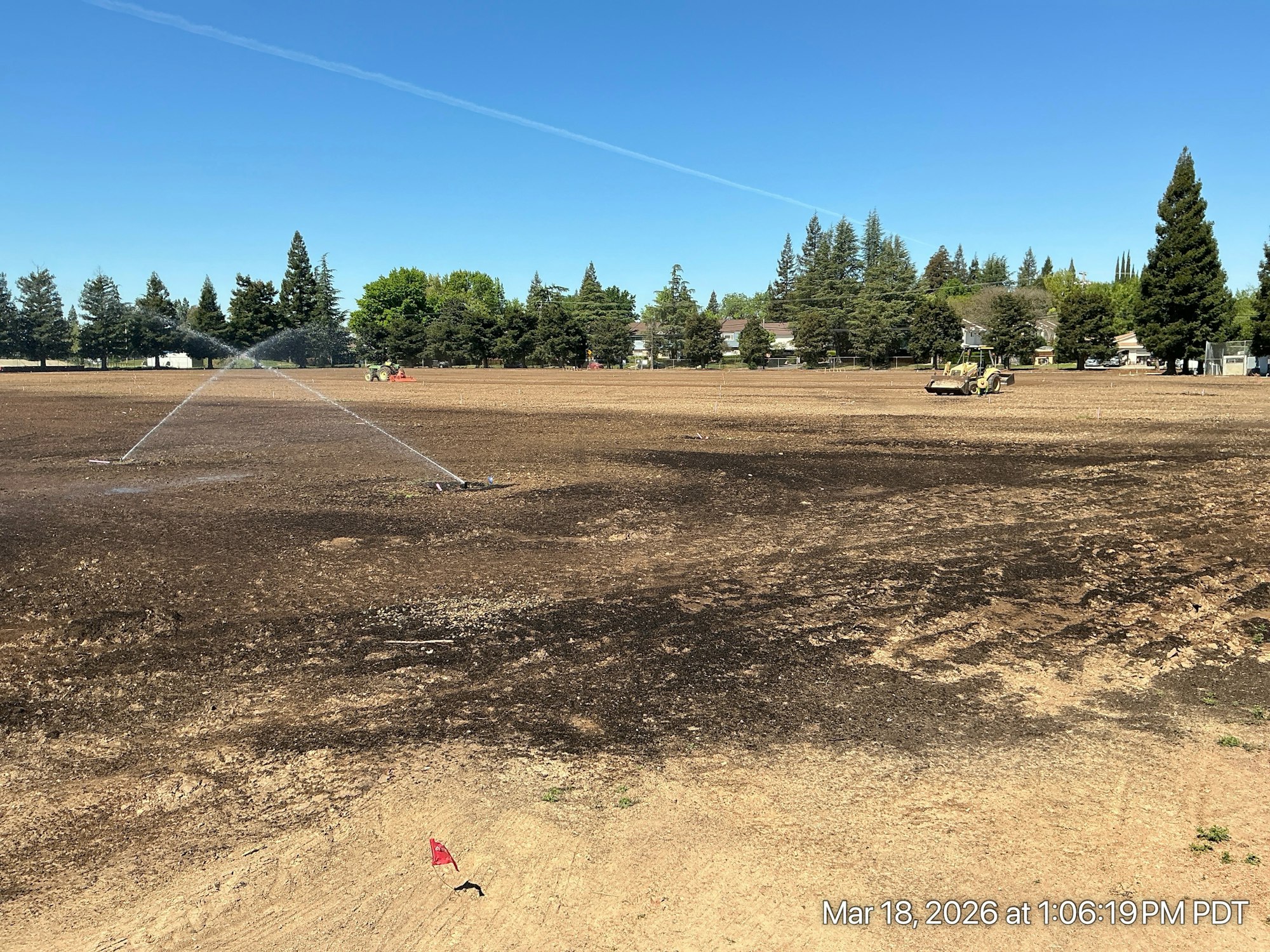 A dry, partially burned field with irrigation systems and a tractor in the background, surrounded by trees and homes.