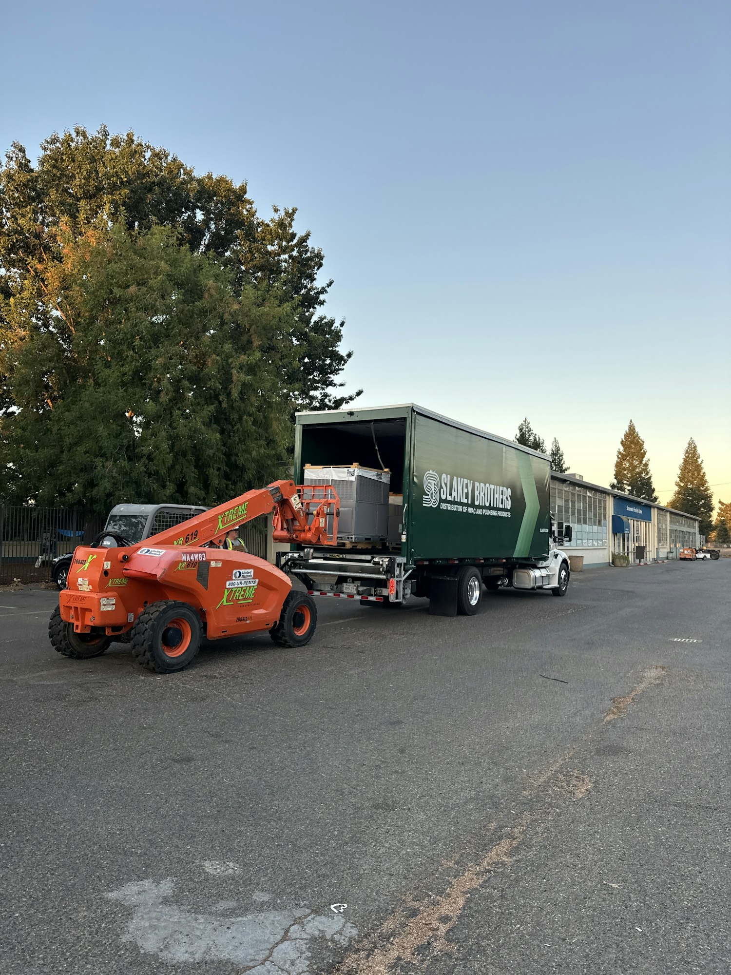 An orange forklift loading a container onto a truck labeled "Slakey Brothers" in an industrial area with trees and buildings.