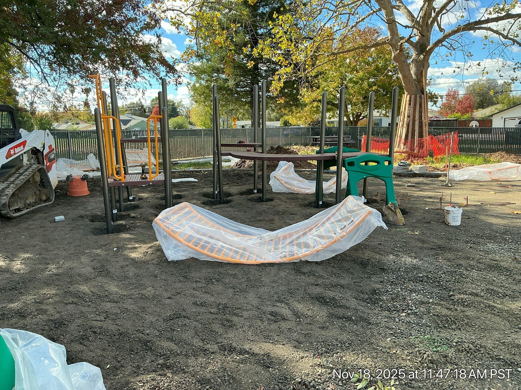 The image shows a playground under construction with partially assembled equipment, machinery, and protective coverings.