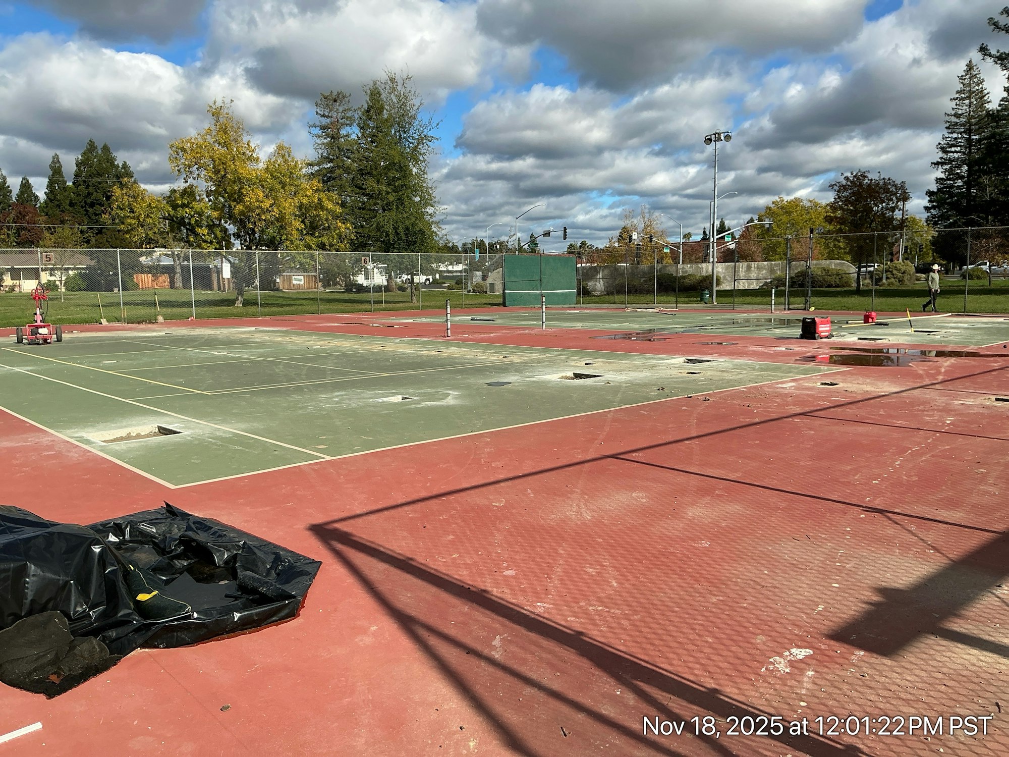 The image shows a set of tennis courts under renovation, with equipment, debris, and cloudy skies visible.