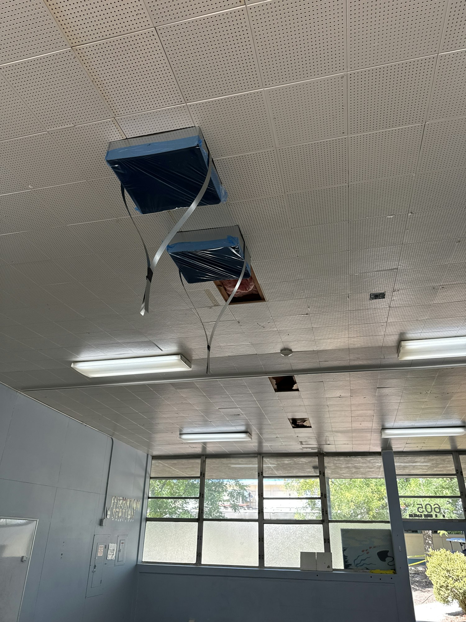 Damaged ceiling with missing tiles, plastic-wrapped sections, and fluorescent lights; large windows below.