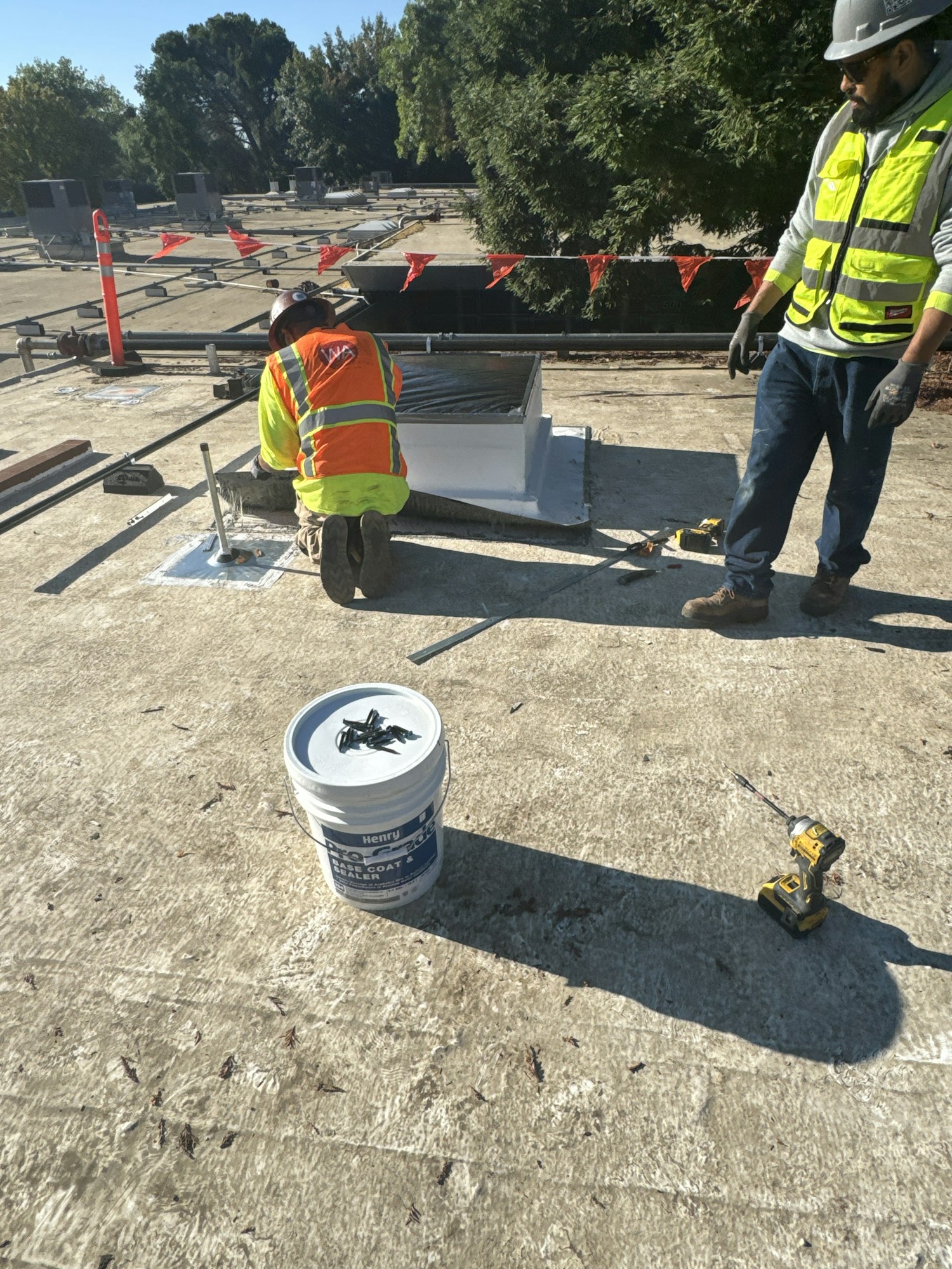 Two workers on a rooftop installing or repairing something, with tools, a bucket, and safety gear.