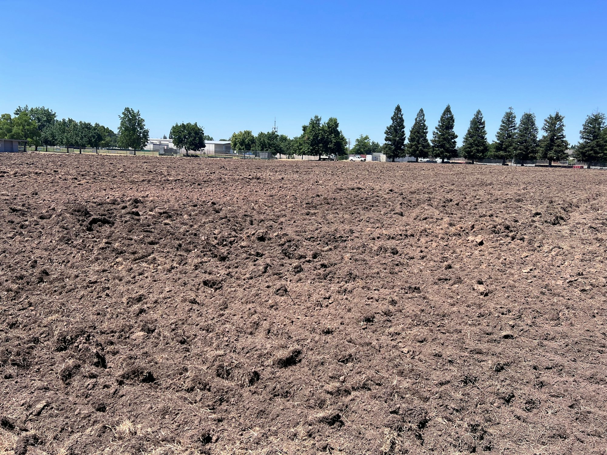 A large, freshly plowed dirt field with trees and buildings in the background under a clear blue sky.