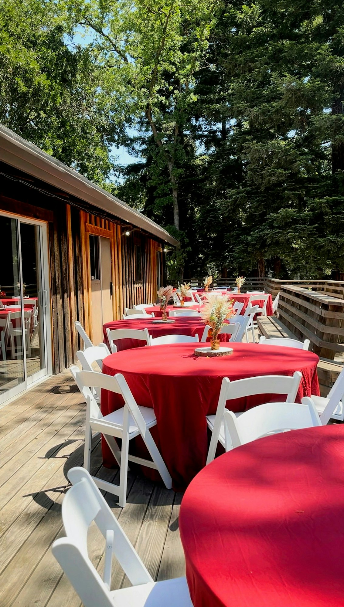 Outdoor wooden deck with round tables covered in red cloth, white chairs, and floral centerpieces; surrounded by trees.