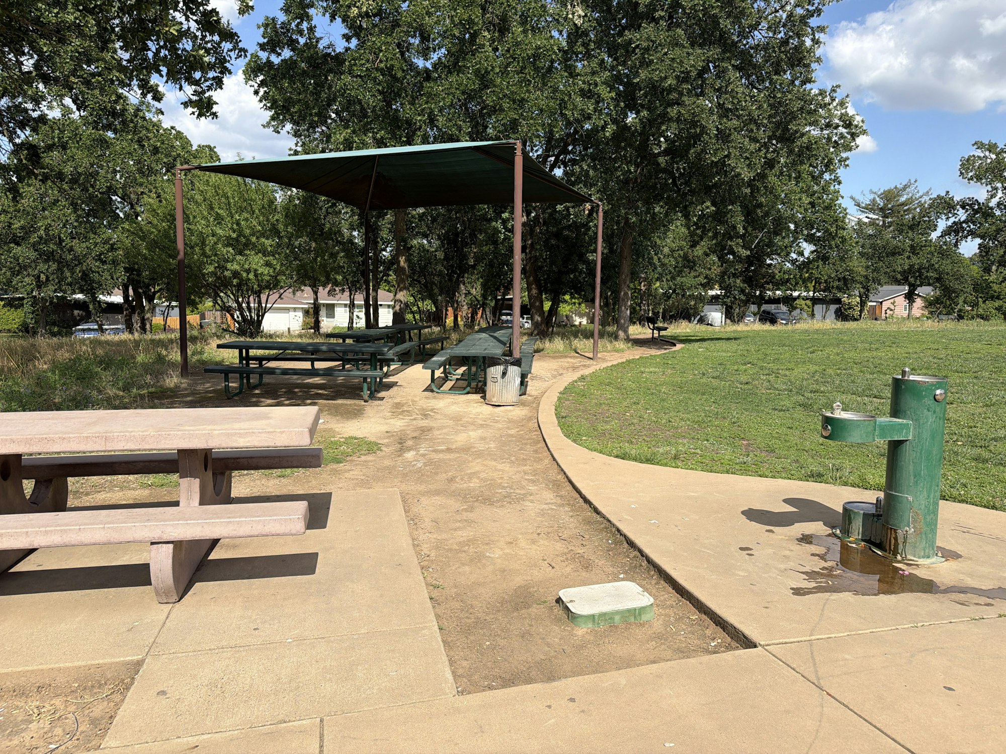 A park area featuring shaded picnic tables, a grassy field, and a water fountain nearby.