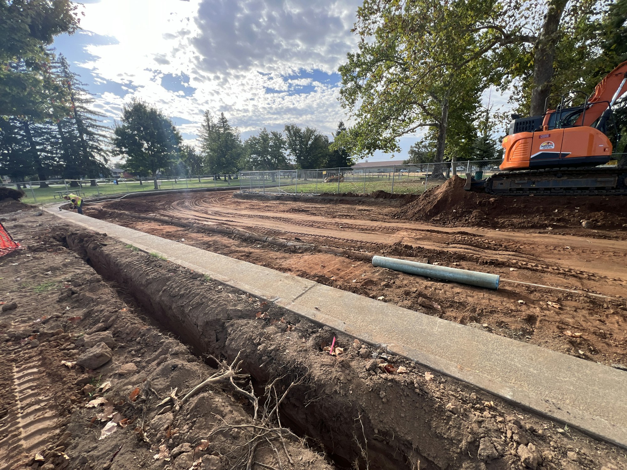 A construction site with a trench, orange excavator, dirt tracks, and a worker. Trees and cloudy sky in the background.