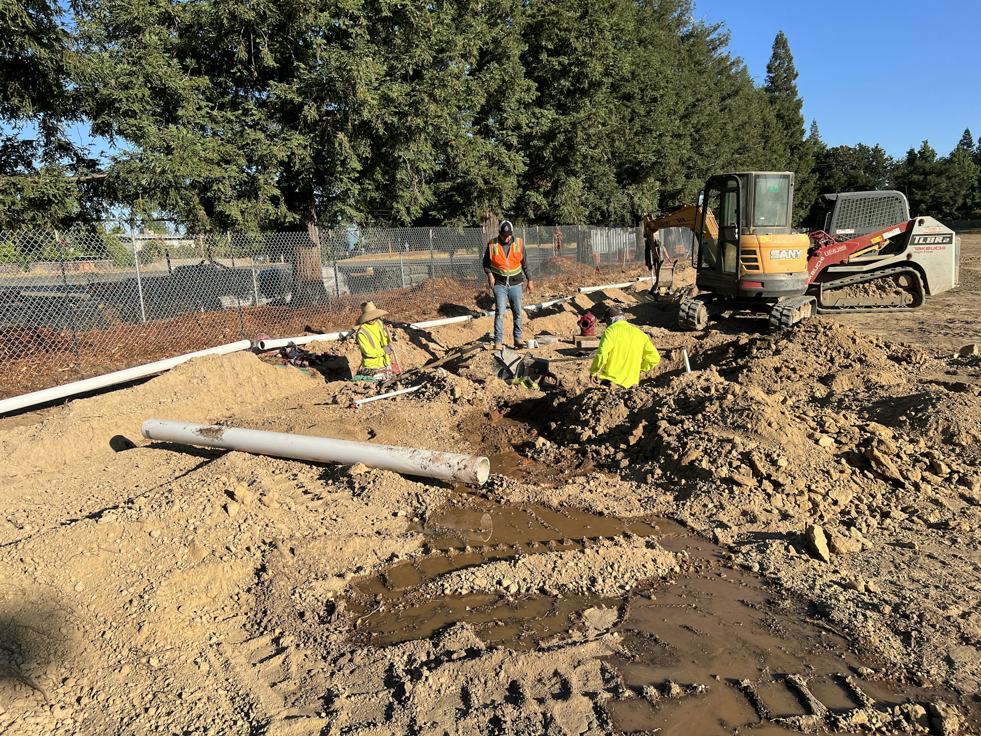 Construction workers installing pipes at a muddy site with machinery and safety gear.
