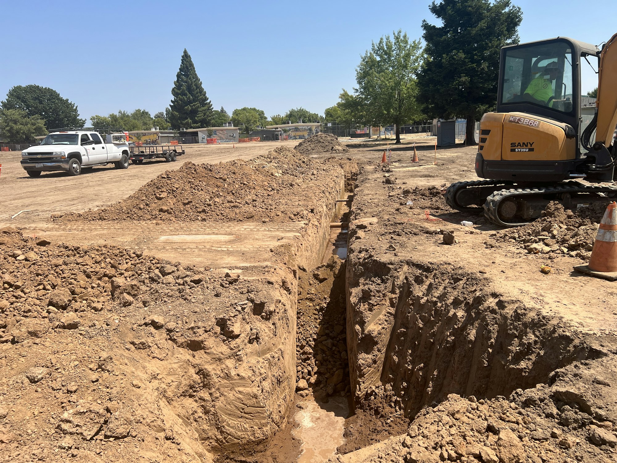 Construction site with a deep trench, an excavator, a white truck, and trees in the background.
