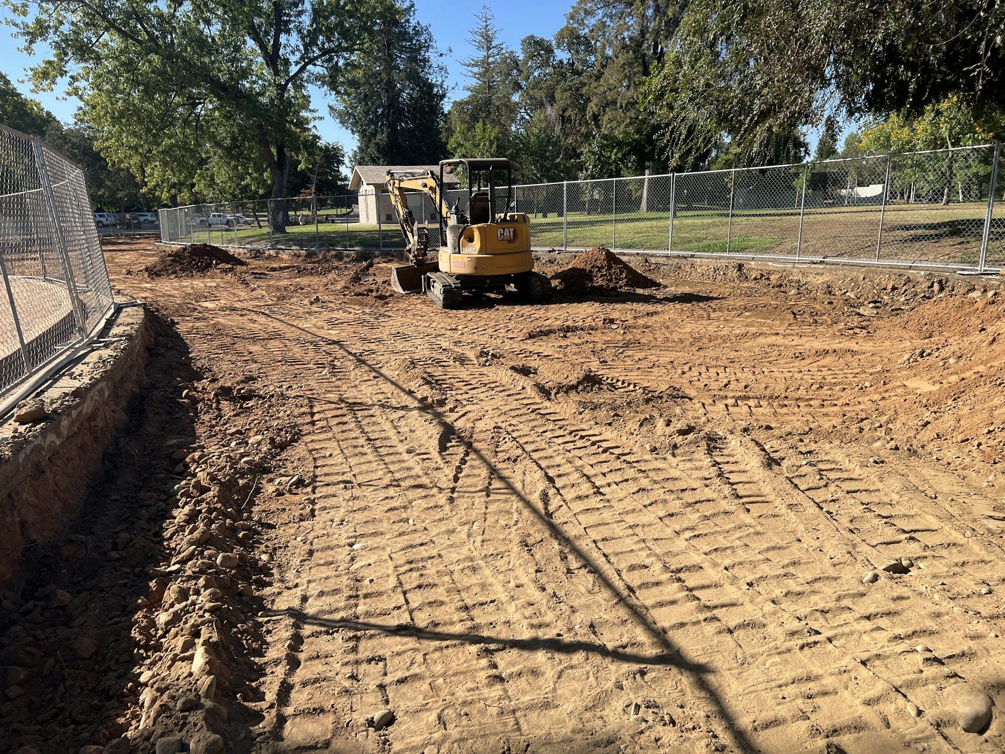 Construction site with an excavator on dirt ground, surrounded by fencing and trees in the background.