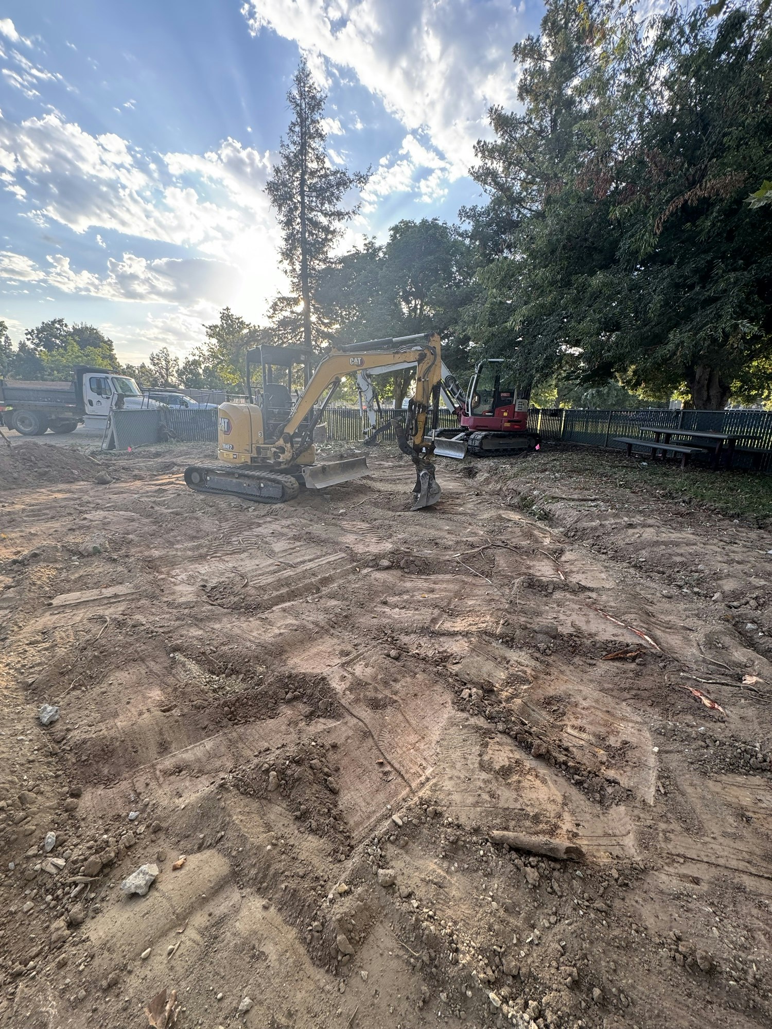 A small excavator on a dirt construction site with trees and a truck in the background.