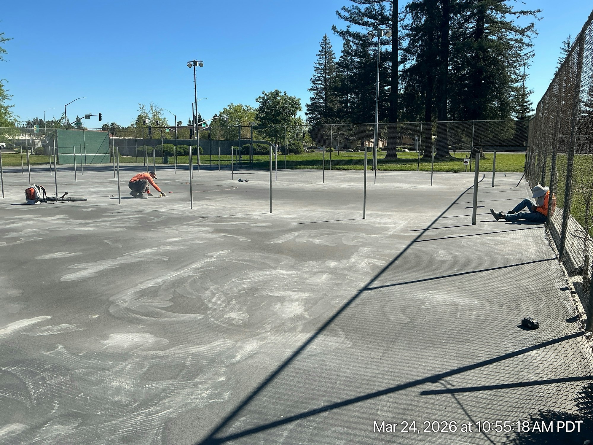 Workers are on a court, possibly preparing for construction, with one sitting and another measuring. Clear blue sky above.