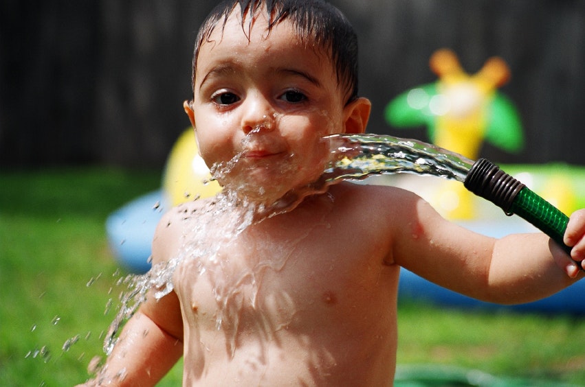 A smiling young child shirtless, playfully splashing water from a hose in a grassy area, with a colorful inflatable toy in the background.