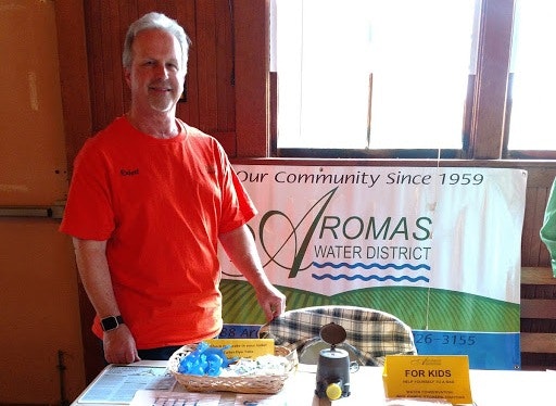 A man in an orange shirt stands at a table with "Aromas Water District" banner, showcasing items for kids and community engagement.
