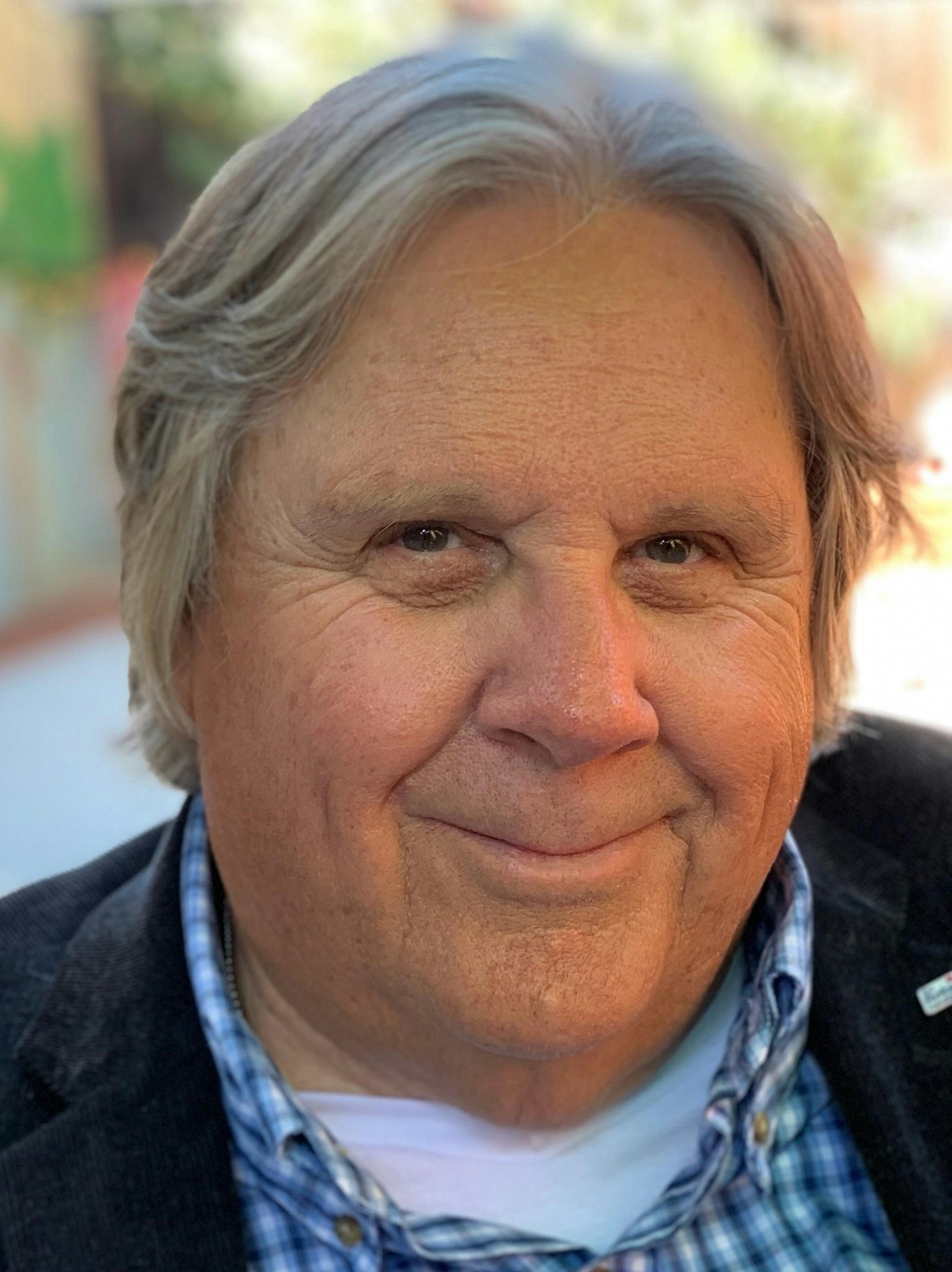 A close-up portrait of a smiling man with gray hair, wearing a plaid shirt and a dark jacket, against a blurred background.