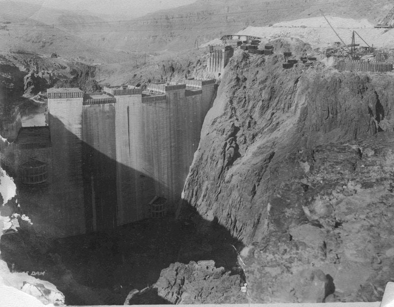The image shows a large dam under construction, surrounded by rocky terrain, with machinery and scaffolding visible.